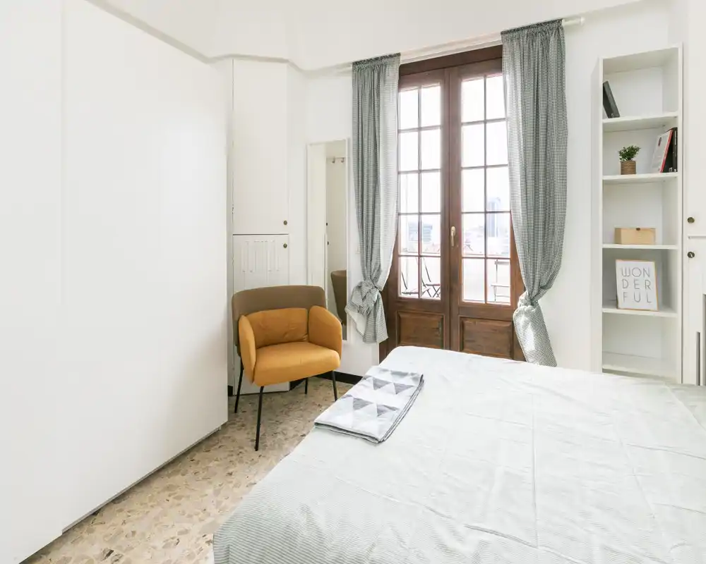 Bright, modern bedroom with a large window and a neatly made bed; soft textiles and a warm accent chair create a welcoming atmosphere.