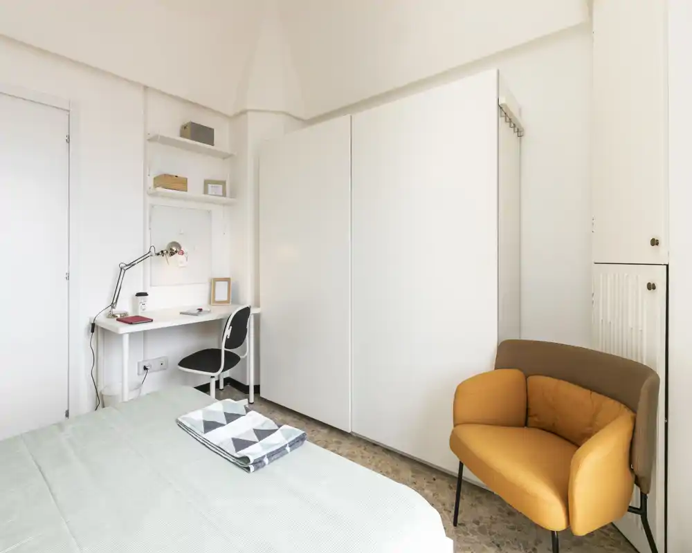 Bright, modern bedroom with a neatly made bed, a compact workspace with desk and lamp, and a warm mustard armchair — ideal for comfortable short or long stays.