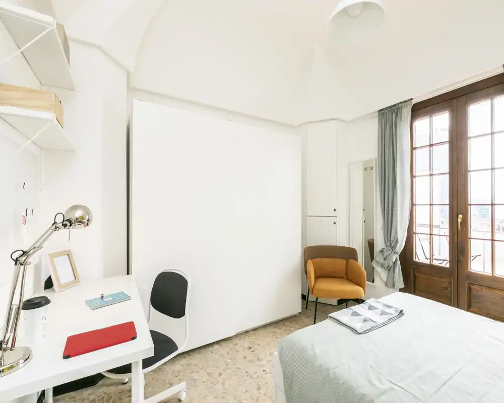 Bright, well-staged bedroom with a comfortable double bed, a small workspace, and a cozy accent chair near French-style glazed doors letting in abundant natural light.