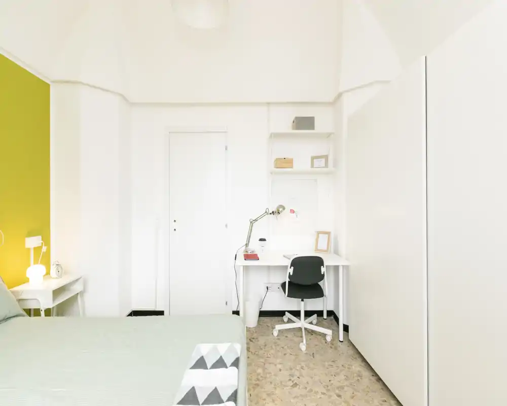 Bright, minimalist bedroom with a visible bed, bedside table and a functional desk area — ideal for a single occupant or student. White walls and a green accent wall create a fresh, airy atmosphere.