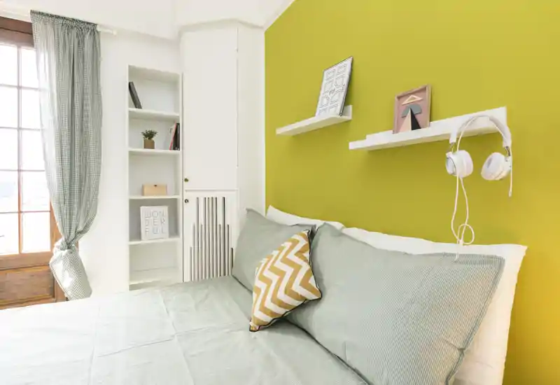 Bright, modern bedroom with a green accent wall, neatly made bed, decorative cushions and wall shelves — ideal for rental listings highlighting a cozy, sunlit sleeping area.