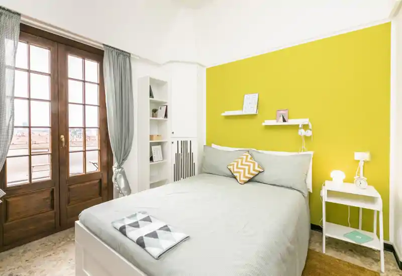 Bright, well-styled bedroom with a double bed against a vivid yellow accent wall, bedside table and large wooden-framed window letting in abundant natural light.