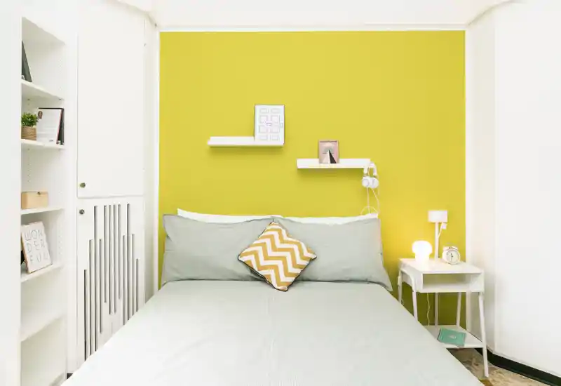 Bright, modern bedroom with a full bed centered against a bold yellow accent wall, a chevron cushion and neat bedside table — styled and ready for guests.