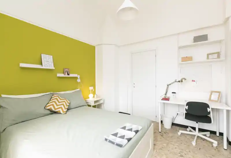 Bright, modern bedroom with a green accent wall, double bed with soft linens, bedside lamp and a small desk workspace.