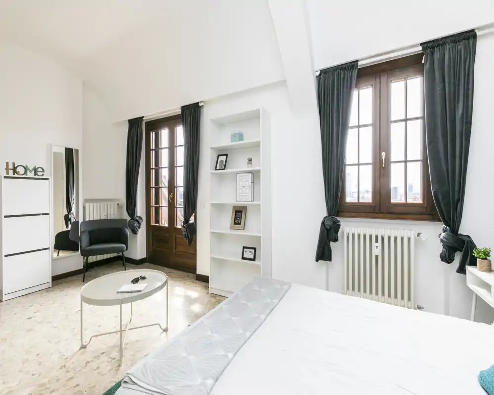 Camera da letto luminosa e moderna con ampie finestre con cornici in legno, letto ben rifatto e arredi minimal in stile scandinavo che rendono l'ambiente arioso e accogliente.