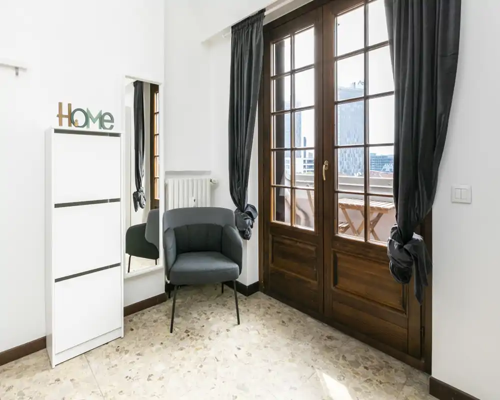 Angolo luminoso del soggiorno con una poltrona comoda, specchio a tutta lunghezza e ampie porte-finestre in legno che si aprono su un piccolo balcone — perfetto come angolo lettura.
