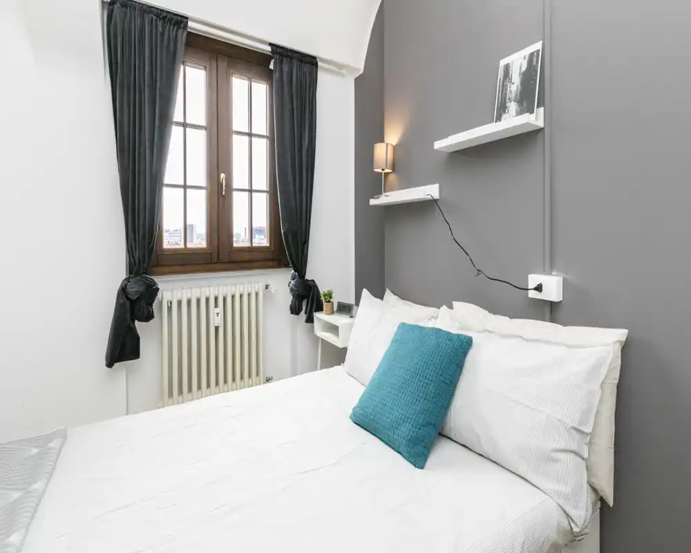 Camera da letto moderna e luminosa con letto matrimoniale rifatto, cuscino turchese e finestra che offre luce naturale e vista sulla città.