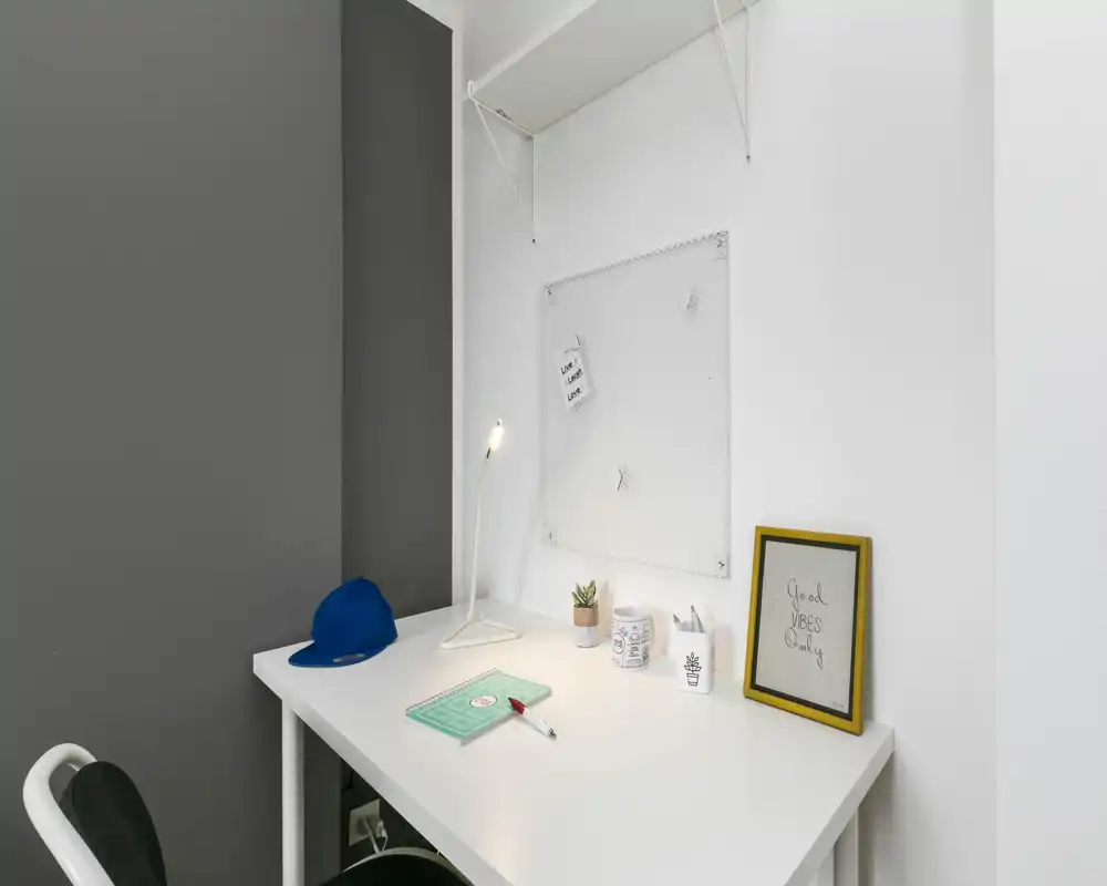 Angolo workspace compatto con scrivania bianca, lampada da tavolo, piccola pianta e cornice decorativa — ideale per studio o lavoro da remoto.
