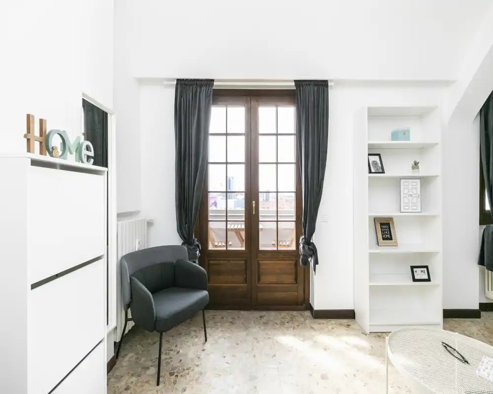 Zona giorno luminosa e minimal con porta finestra a vetri che apre sul balcone, poltrona moderna e mensole aperte — perfetta per un appartamento urbano.