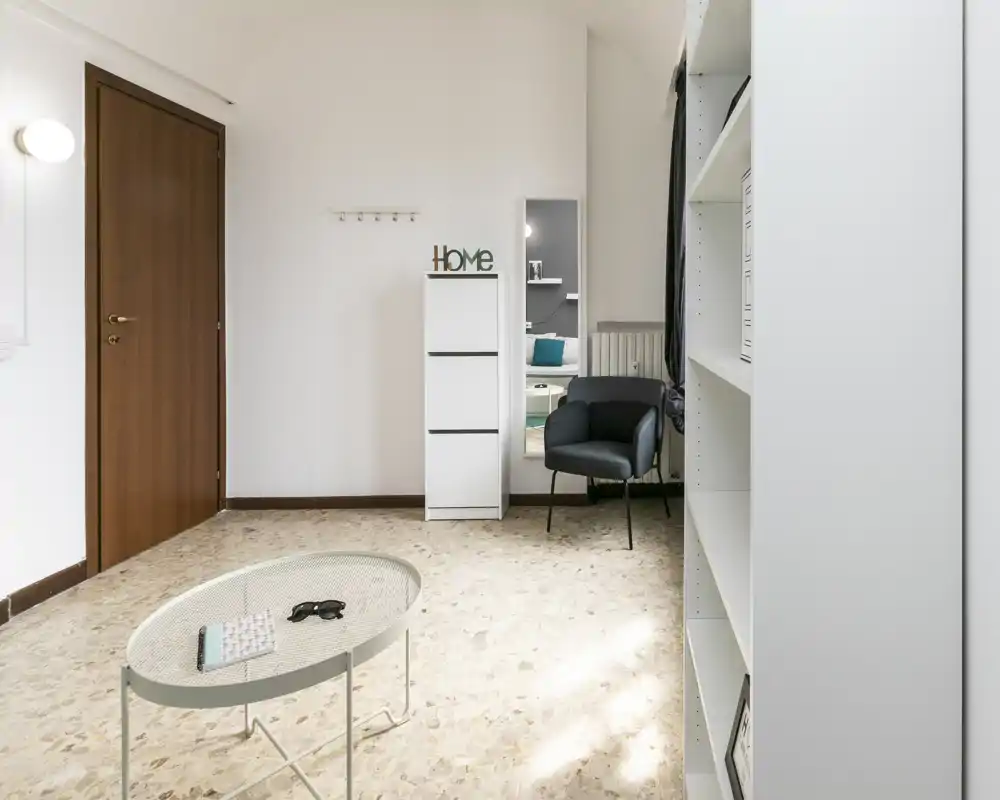 Zona giorno luminosa e minimal con una poltroncina, tavolino e scaffale; toni neutri e uno specchio che amplifica lo spazio.