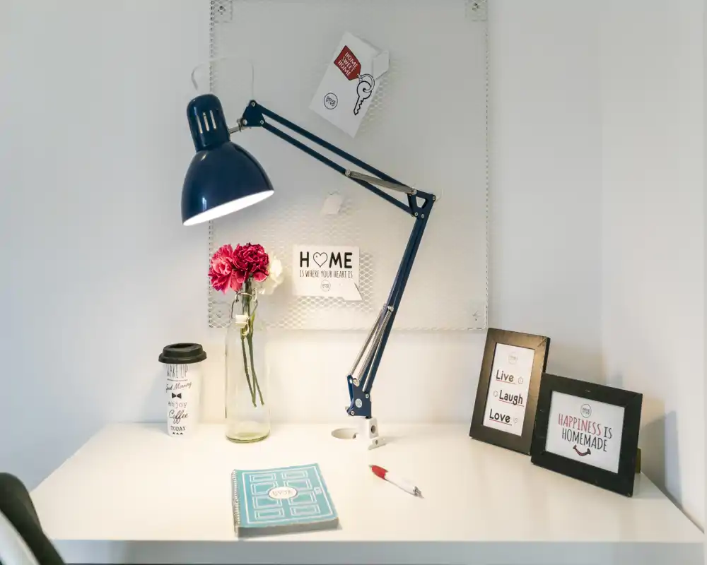 A bright, neatly styled bedside or small workspace with a lamp, framed prints, a notebook and fresh flowers—clean, welcoming and well-lit.