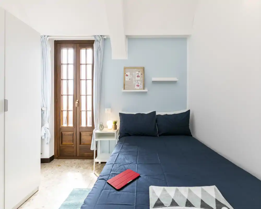 Bright, tidy bedroom with a double bed dressed in navy linens, a bedside table with lamp, and natural light from wooden French doors.