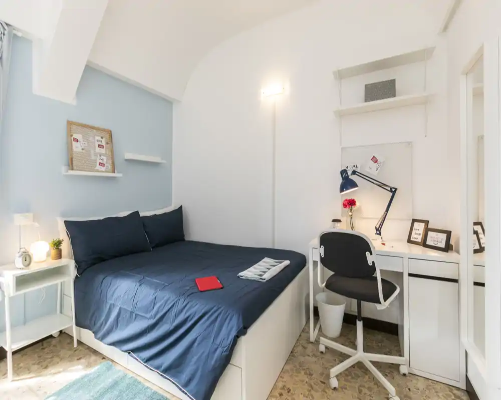 Bright, well-styled bedroom with a navy bedspread, bedside table and a compact white desk with lamp — ideal for a single occupant or a cozy rental.