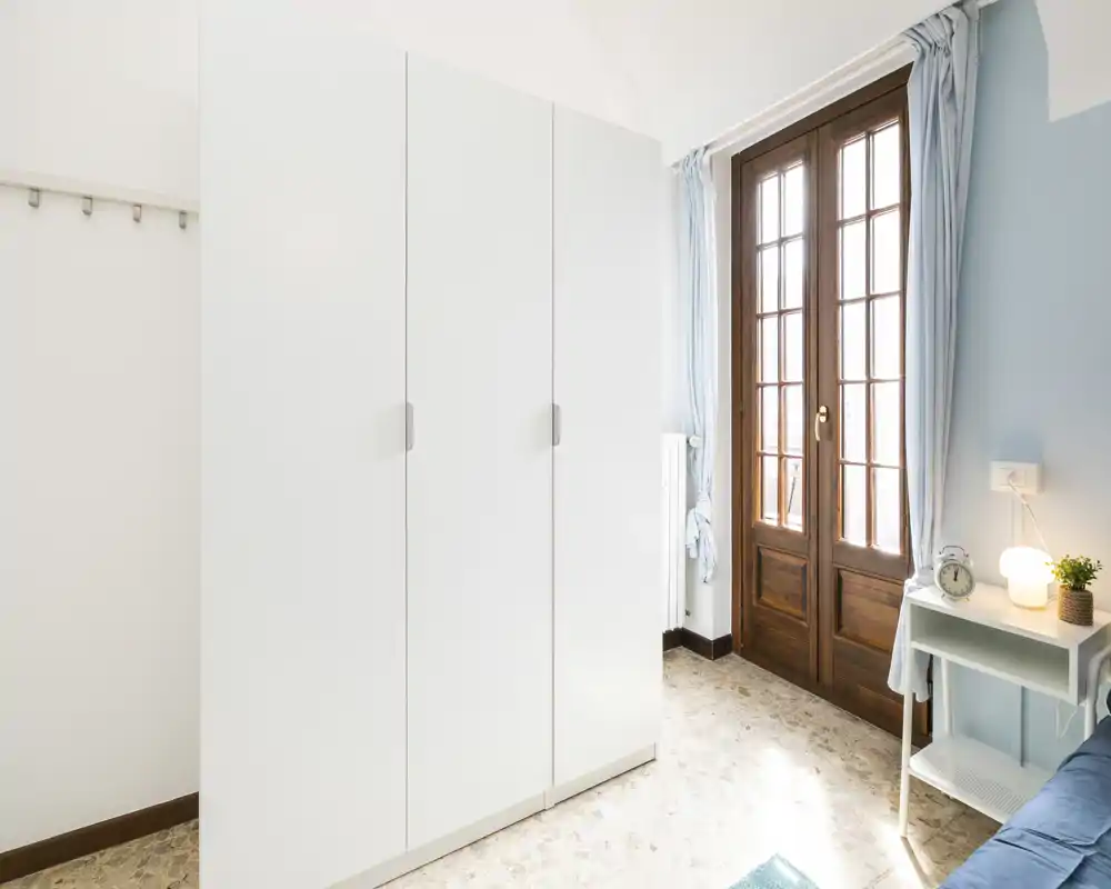 Small, bright bedroom corner with a large white wardrobe, wooden-framed glass door letting in natural light, and a bedside table with lamp and alarm clock.