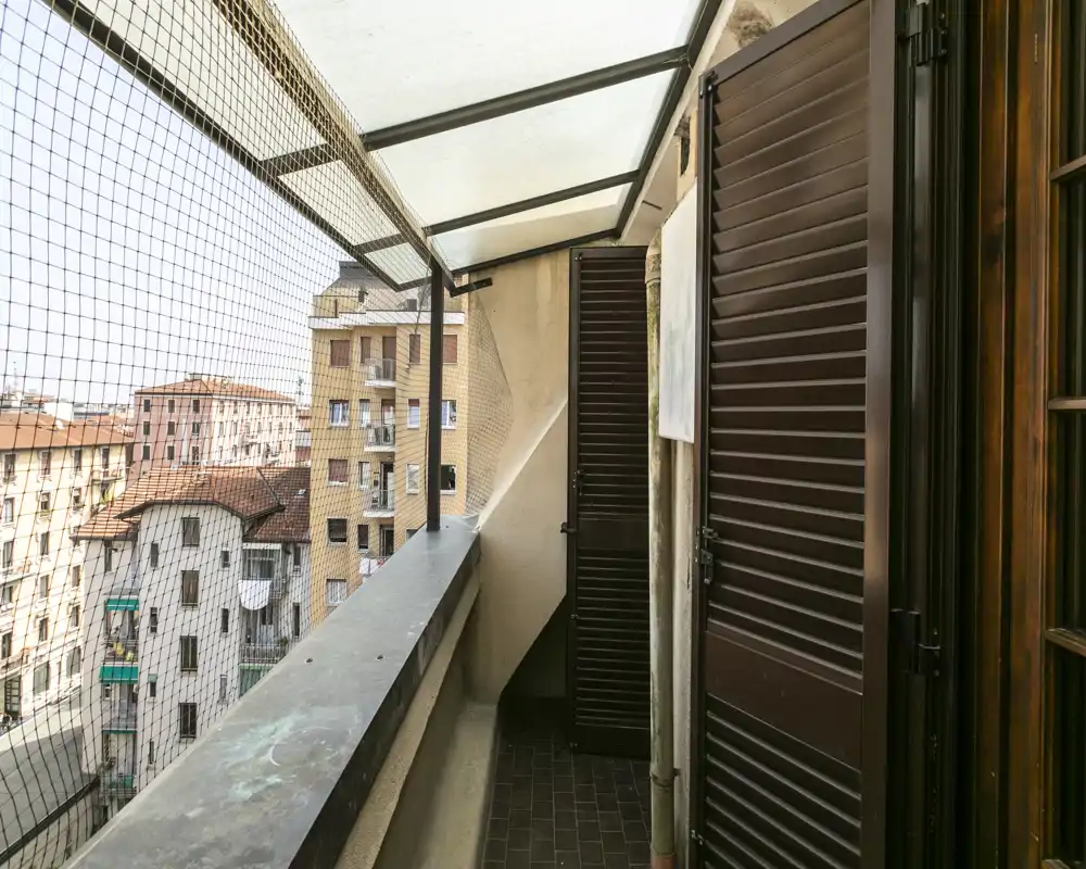 Narrow covered balcony with protective mesh, city view and shuttered doors leading inside.