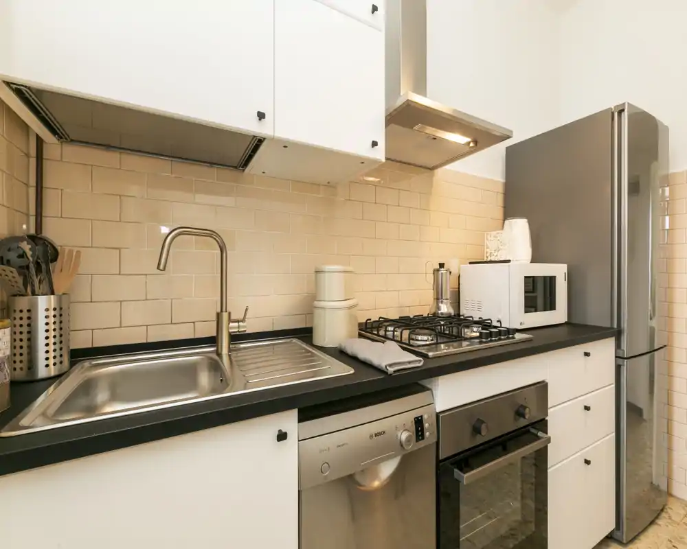 Modern, compact kitchen with stainless-steel sink, gas hob, built-in oven, microwave and tall refrigerator. Light subway-tile backsplash and black countertop give a clean, contemporary look—ideal for rental listing headlines.
