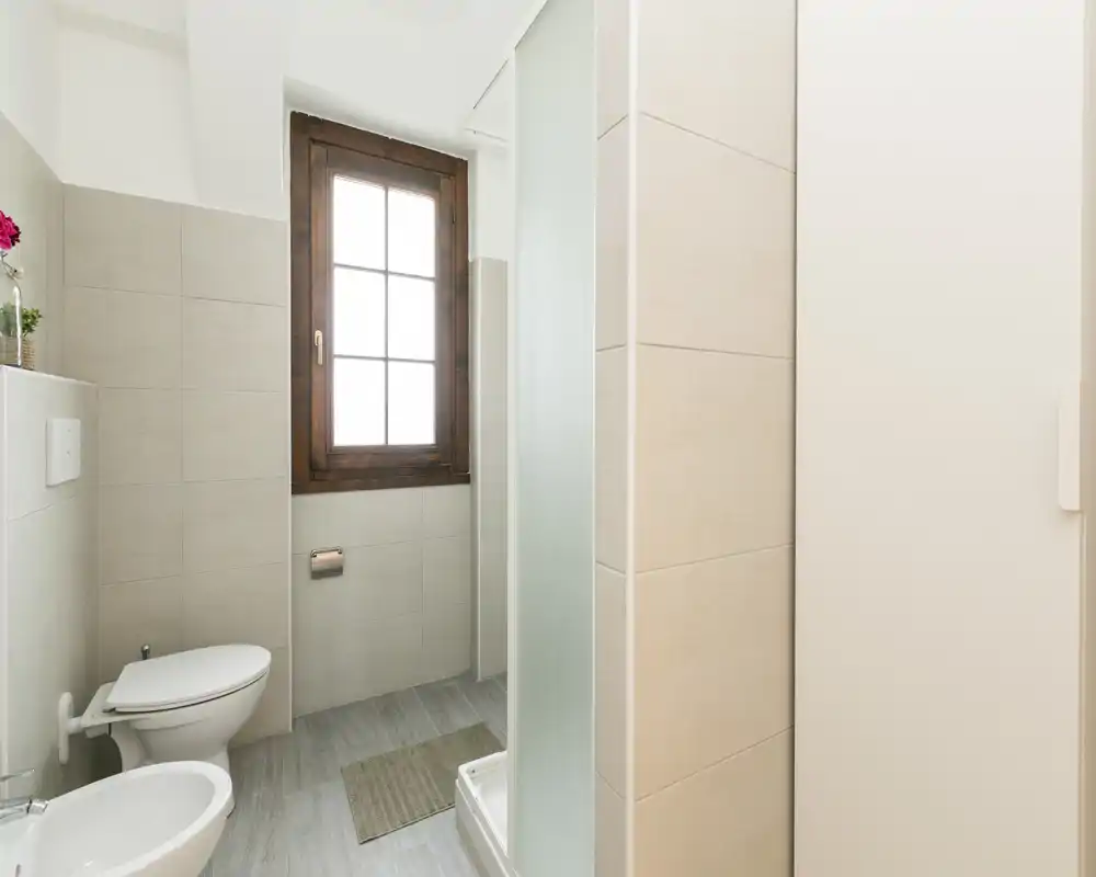 Bright, modern bathroom with a toilet, bidet, shower stall and a wooden-framed window; neutral tiles and pale flooring create a clean, airy feel.