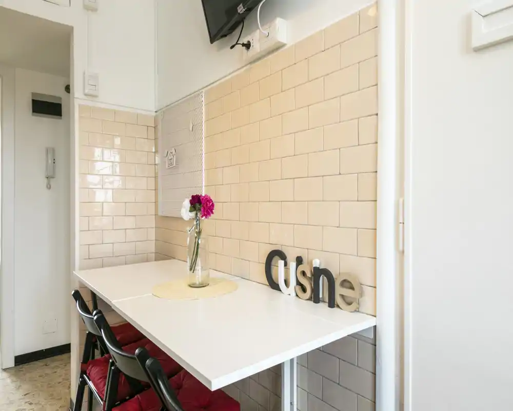 Compact kitchen breakfast nook with a white folding table, three chairs with red cushions and decorative flowers on the table — bright, tidy space ideal for casual meals.