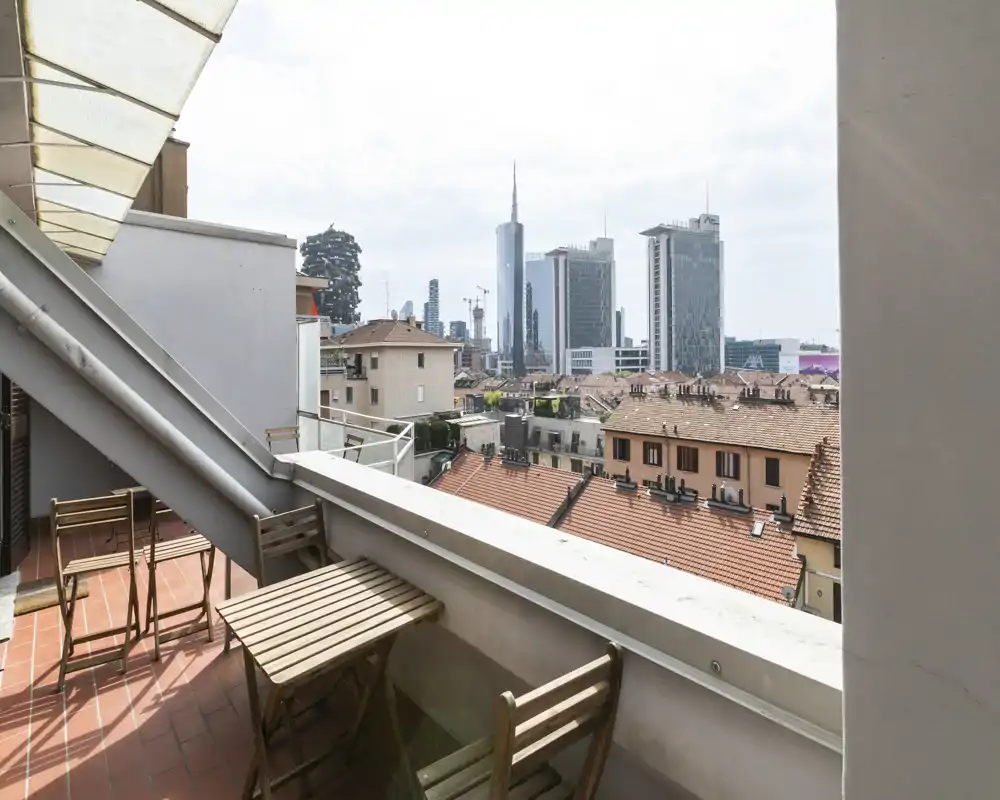 Sunny balcony with a small wooden table and folding chairs overlooking a city skyline with modern high-rises — ideal for morning coffee or relaxing outdoors.