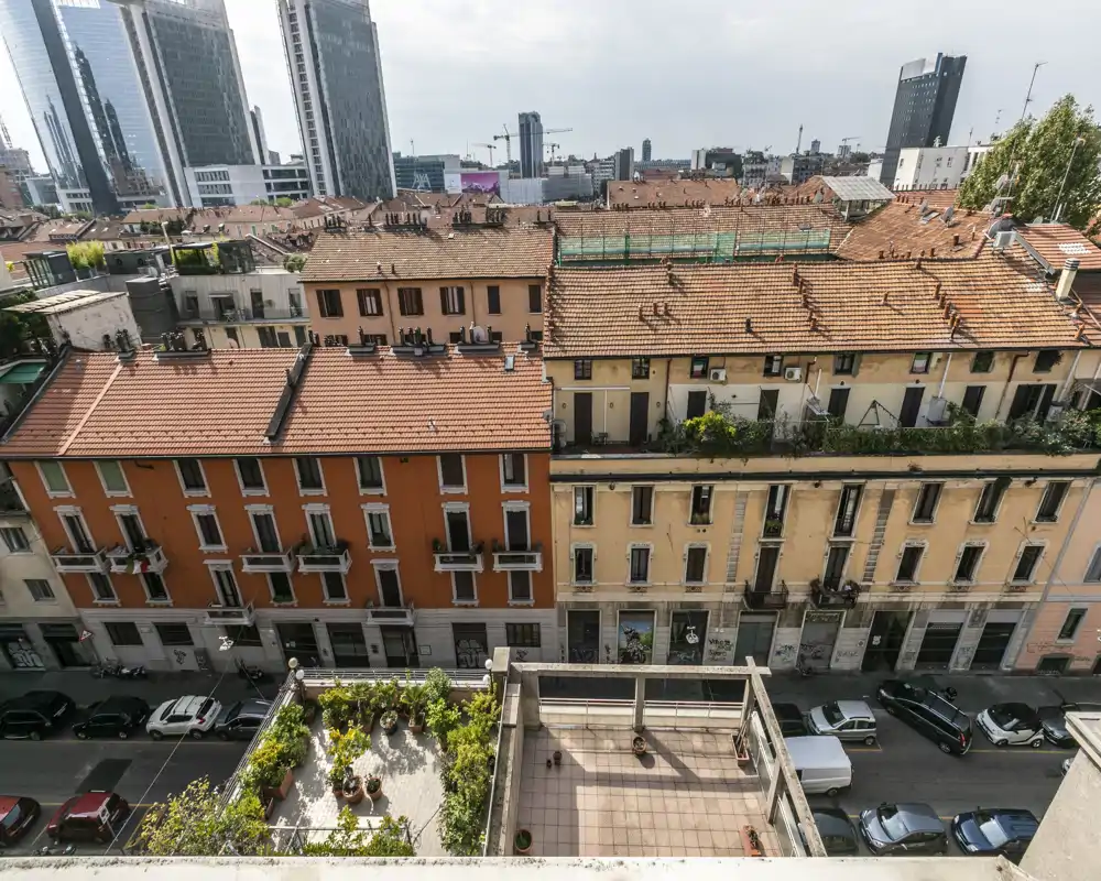 City-facing outdoor terraces and balconies seen from above, with potted plants and tiled patios overlooking a street and neighboring rooftops.