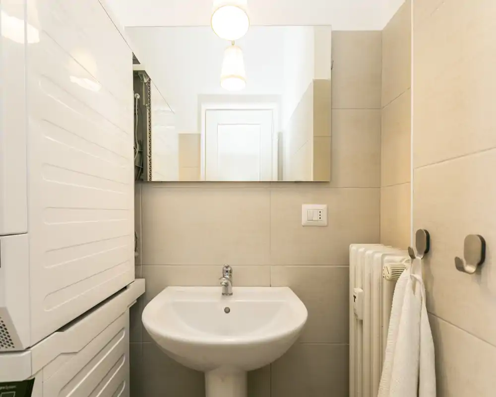 Compact, well-lit bathroom with a pedestal sink, large mirror and stacked laundry appliances visible at left. Neutral tiles and simple fixtures create a clean, functional space.