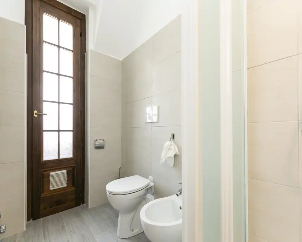 Bright, modern bathroom with a toilet and bidet, light tiles and a wooden framed frosted-glass door providing natural light.