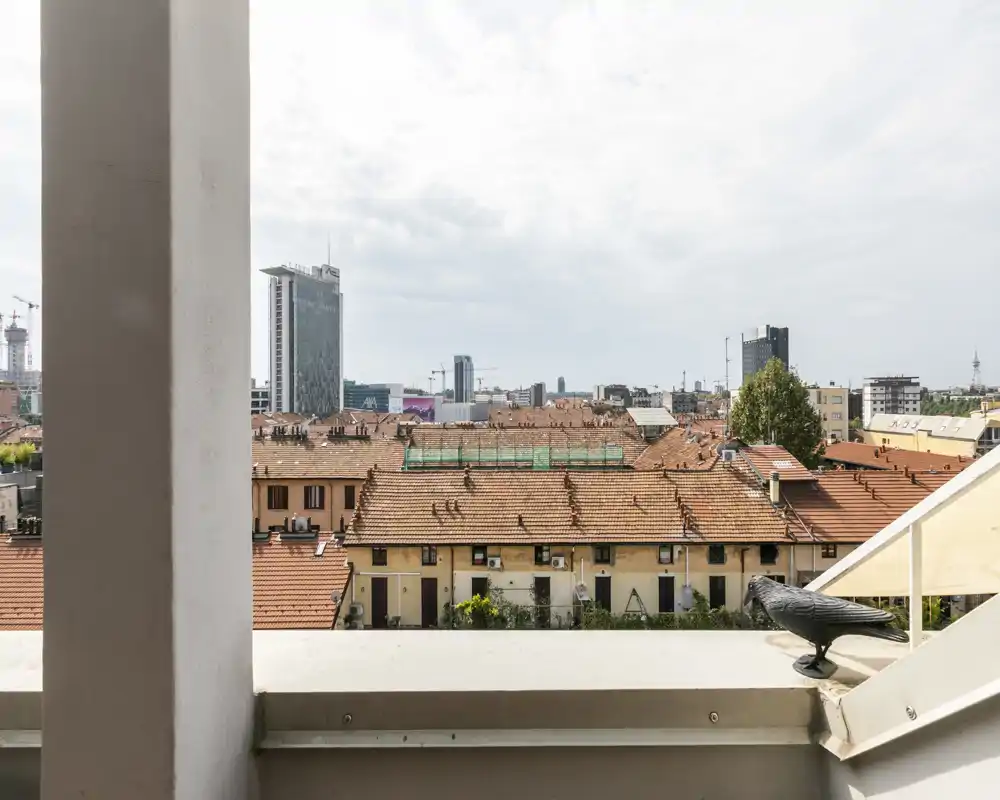 Sunny balcony view over terracotta rooftops and a distant city skyline, with a decorative bird on the railing and clear urban perspective.