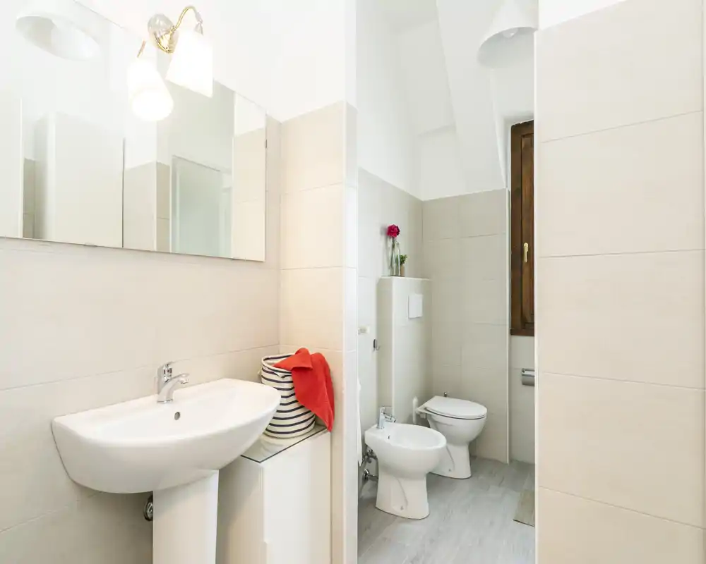 Bright, modern bathroom with a pedestal sink, bidet and toilet, neutral tiles and a wooden-framed window.