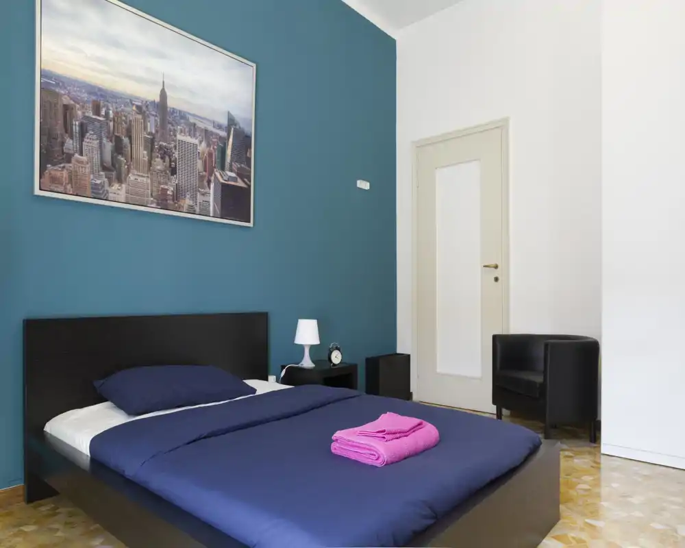 Camera da letto moderna e luminosa con letto matrimoniale basso con biancheria blu navy, comodino con lampada e sveglia e una poltrona nera. La parete color petrolio e il quadro della città creano un’atmosfera elegante e accogliente ideale per gli annunci.