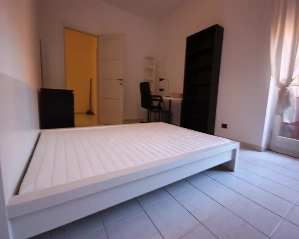 Camera da letto luminosa e minimale con un ampio letto bianco a piattaforma in primo piano, pavimento piastrellato e una piccola zona scrivania: perfetta per un annuncio moderno.