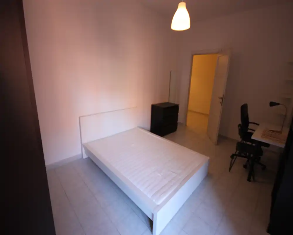 Camera da letto minimal con struttura letto matrimoniale bianca e materasso, un piccolo comò e una scrivania con sedia. Pavimento piastrellato e pareti neutre creano uno spazio semplice e luminoso adatto a un annuncio.