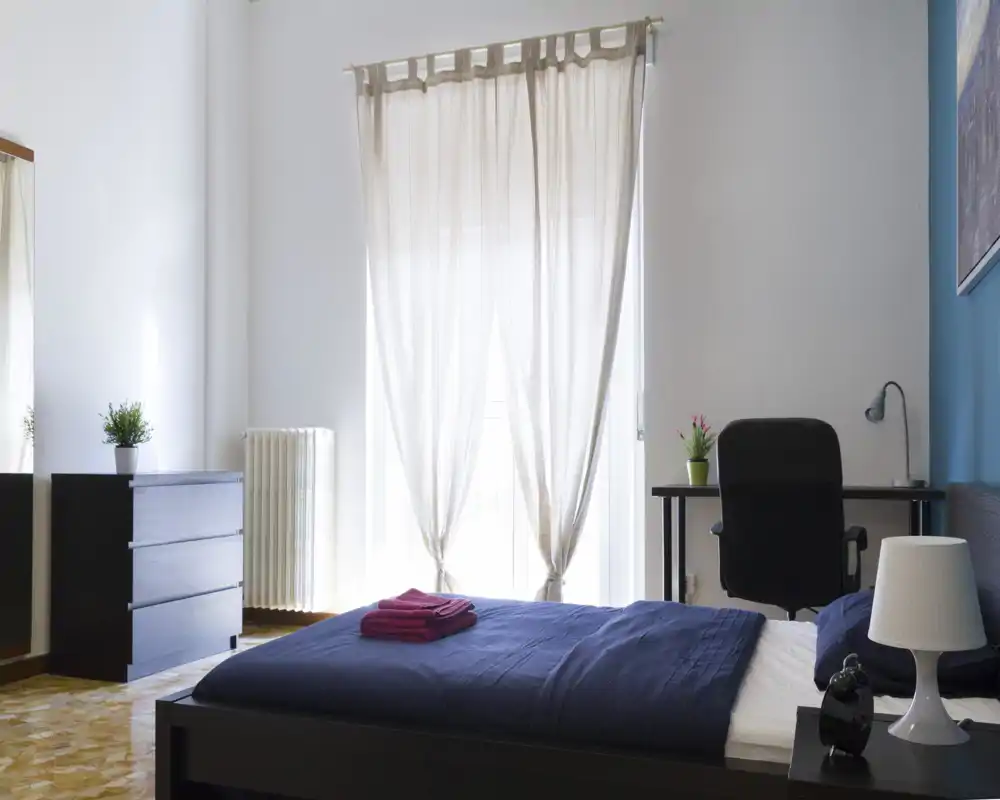 Camera da letto moderna e luminosa con letto ben rifatto, scrivania e comò; luce naturale morbida attraverso le tende trasparenti crea un'atmosfera accogliente.