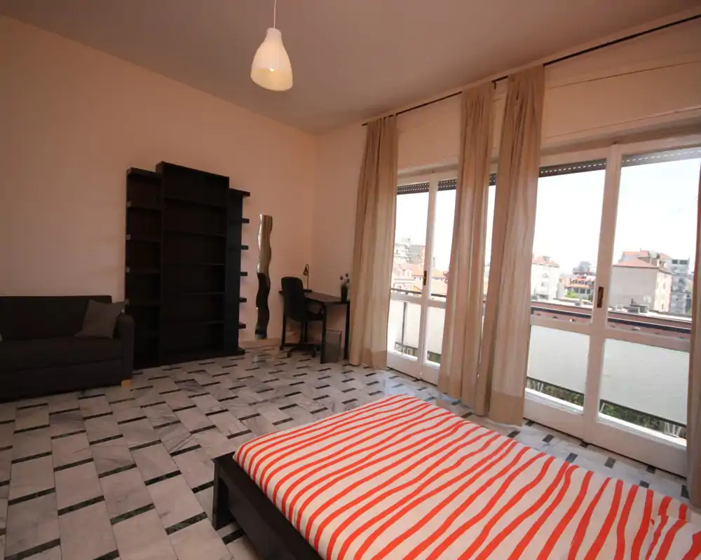 Amplio dormitorio con cama baja de madera en primer plano, grandes ventanales que aportan abundante luz natural, sofá y zona de escritorio, y cortinas neutras para privacidad.