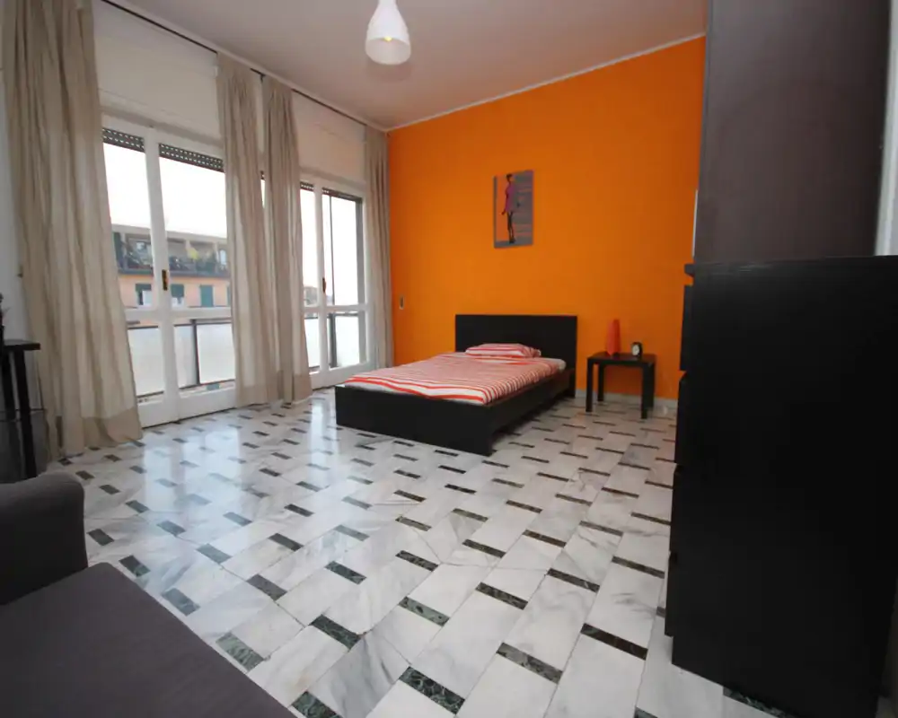 Dormitorio luminoso y minimalista con una cama baja negra frente a una pared naranja llamativa y grandes ventanales que proporcionan mucha luz natural.