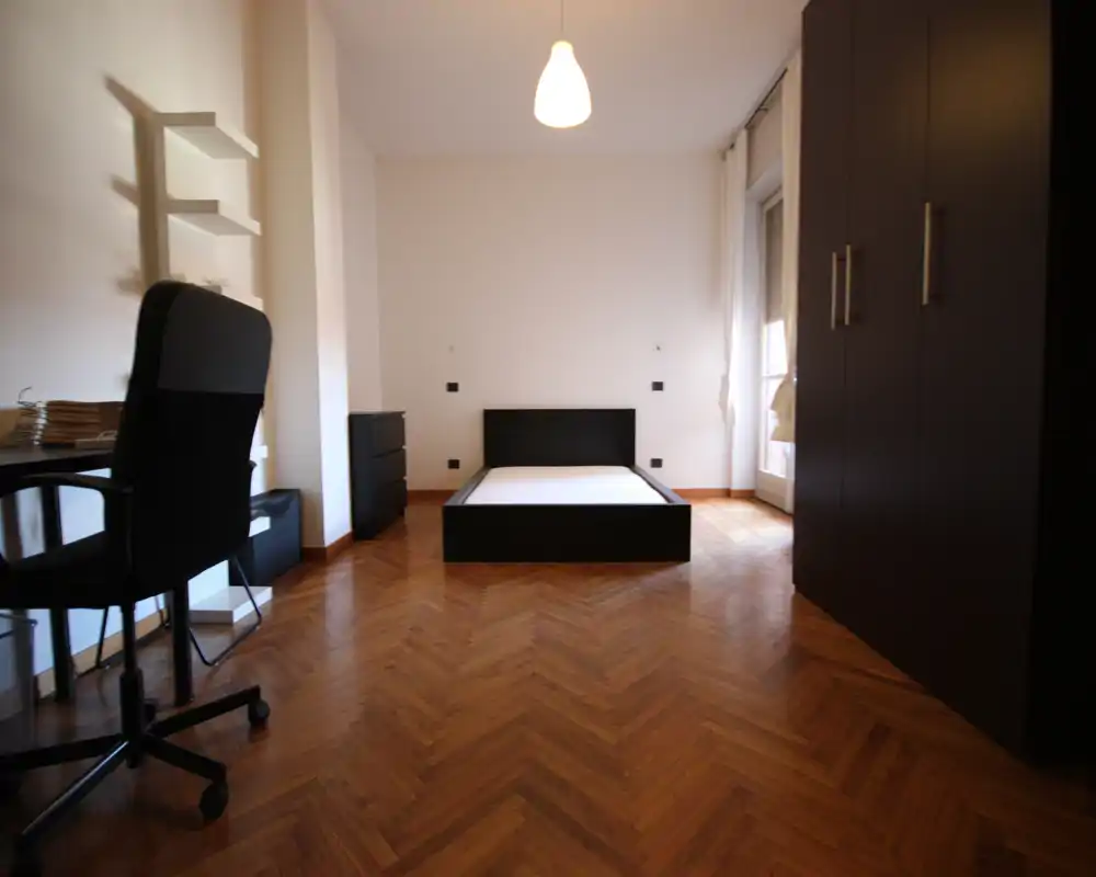 Dormitorio minimalista y moderno con una estructura de cama baja negra centrada contra una pared lisa, gran armario a la derecha, escritorio y silla a la izquierda y suelo de madera en espiga. La luz natural aporta una sensación limpia y luminosa, ideal para un anuncio de alquiler.