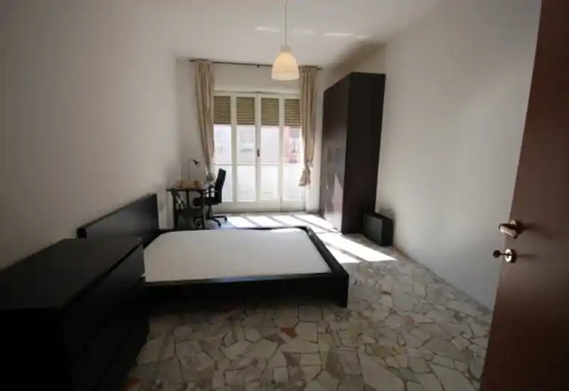 Camera da letto luminosa e semplicemente arredata con letto matrimoniale in cornice, comò, armadio e una scrivania vicino a una grande finestra che offre molta luce naturale.