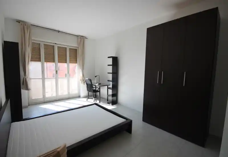 Camera da letto luminosa e minimal con ampia finestra, letto matrimoniale basso, grande armadio scuro e una piccola scrivania vicino alla finestra — spazio semplice e ben illuminato, ideale per un annuncio.