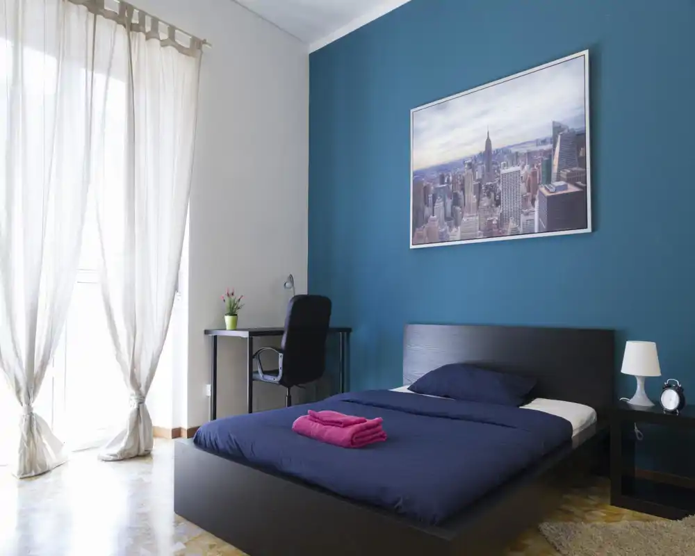 Camera da letto moderna e luminosa con letto basso in legno scuro e biancheria blu navy, comodino con lampada, scrivania con sedia e una grande finestra con tende leggere che illuminano la stanza.