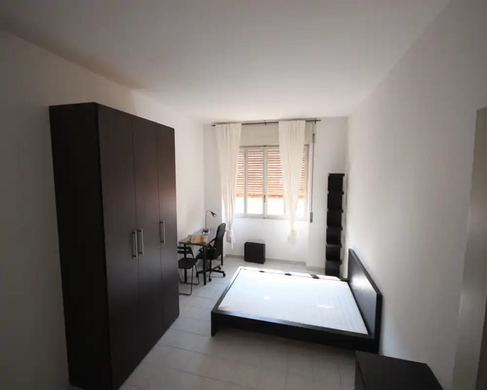 Camera da letto luminosa e minimal con un letto basso, ampio armadio e una piccola scrivania vicino alla finestra. La luce naturale riempie l'ambiente e l'arredamento neutro crea uno spazio calmo e funzionale.