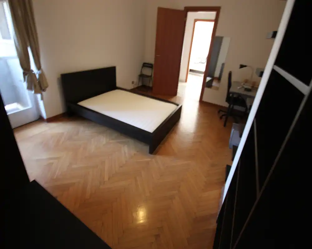 Bright, minimally furnished bedroom with a large double bed frame on chevron parquet flooring and a small desk near the doorway.