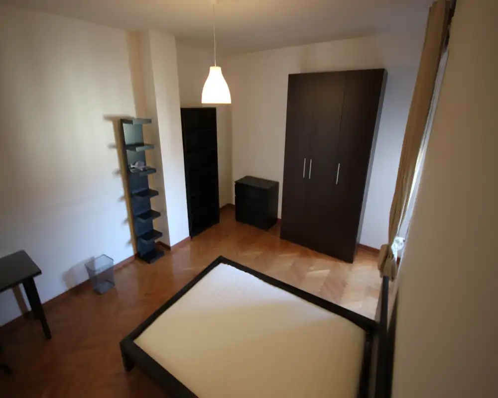 Spacious bedroom with a visible double bed frame, dark wardrobe and matching bedside storage. Natural light from the window highlights the parquet flooring and the room’s clean, minimalist style.