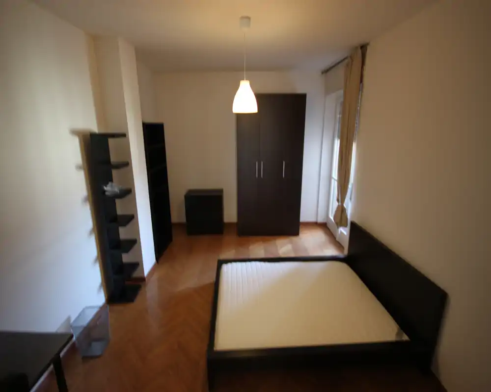 Simple furnished bedroom with a double bed frame, wardrobe and shelves; natural light from a window and a central pendant lamp create a bright, minimal space.