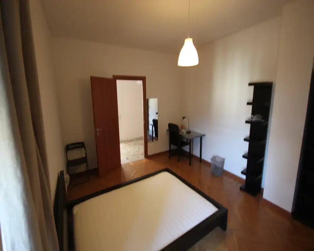 Bright, simply furnished bedroom with a double bed frame, desk and shelving; hardwood floors and natural light from the left window create a neat, functional space.