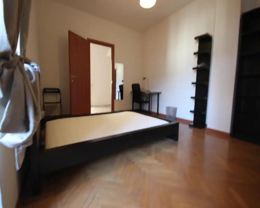Bright, simply furnished bedroom with a low wooden bed frame, parquet floor and shelving. A desk and chair sit against the far wall; natural light comes from the left window.