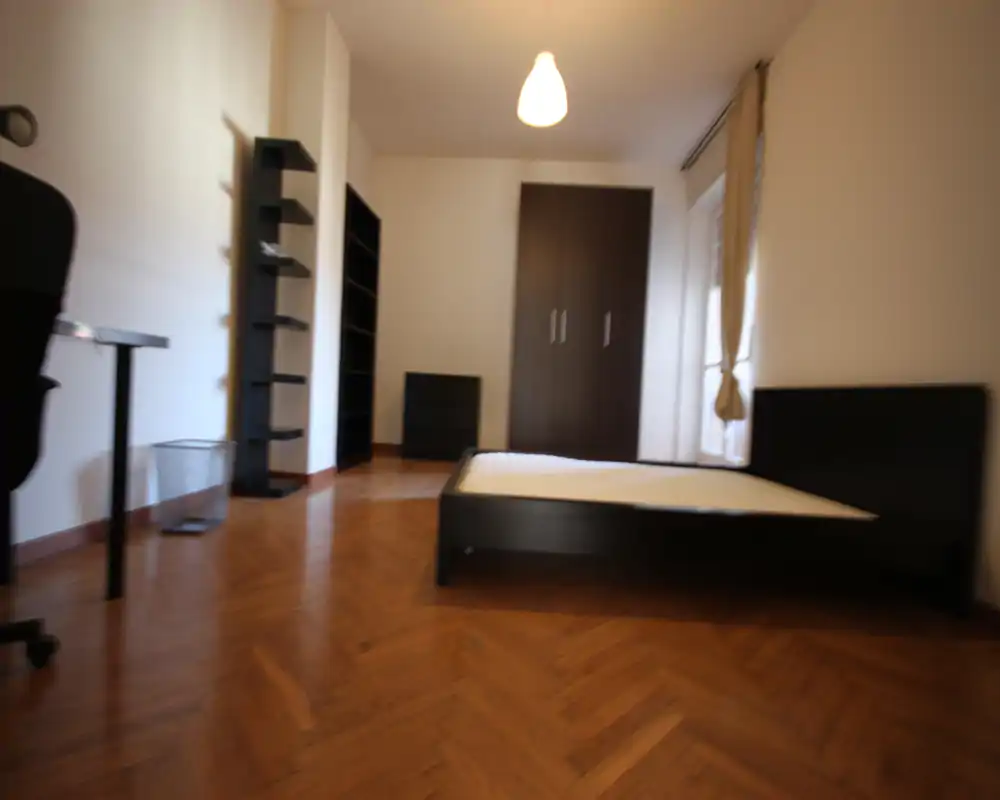 Bright, simply furnished bedroom with a low black bed frame, wardrobe and shelving; wooden parquet floor and a window providing natural light.