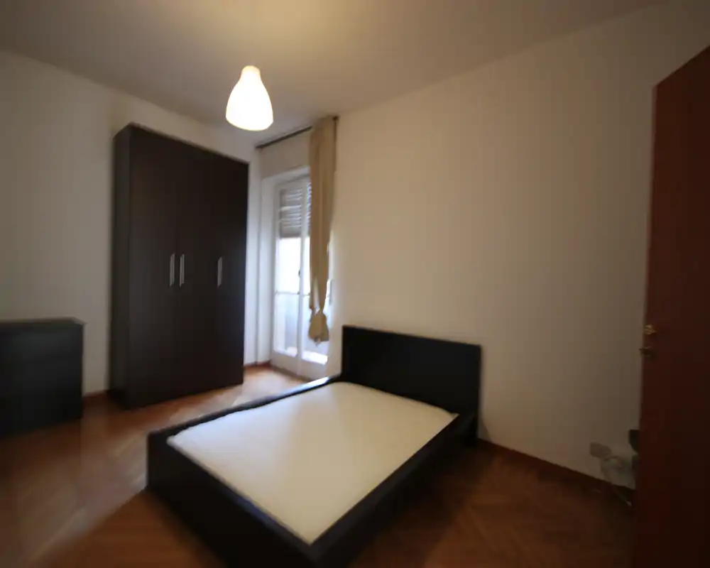 Simple bedroom with a double bed frame and mattress, a wardrobe and a chest of drawers; natural light from a balcony door provides a bright atmosphere.