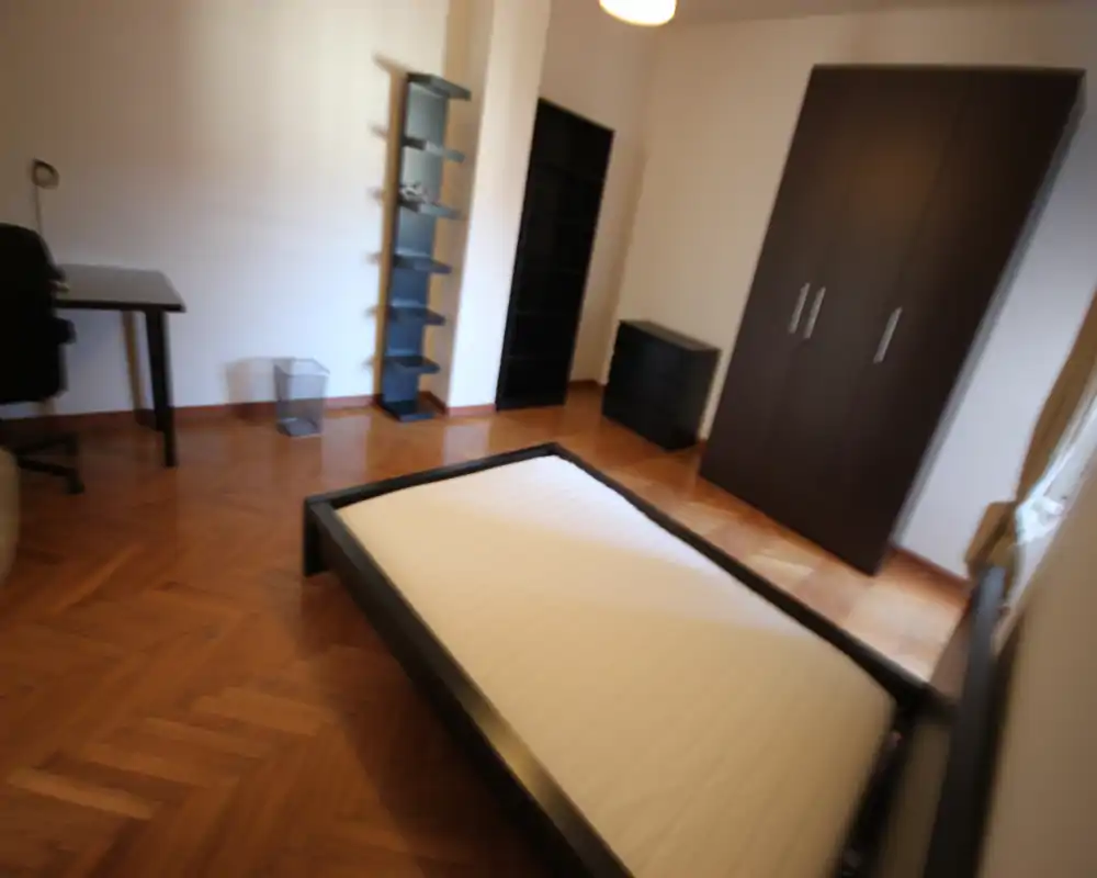Bright bedroom with a dark wood bed frame and mattress, matching wardrobe and shelving, and parquet flooring. The room is simply furnished and receives natural light from a side window.