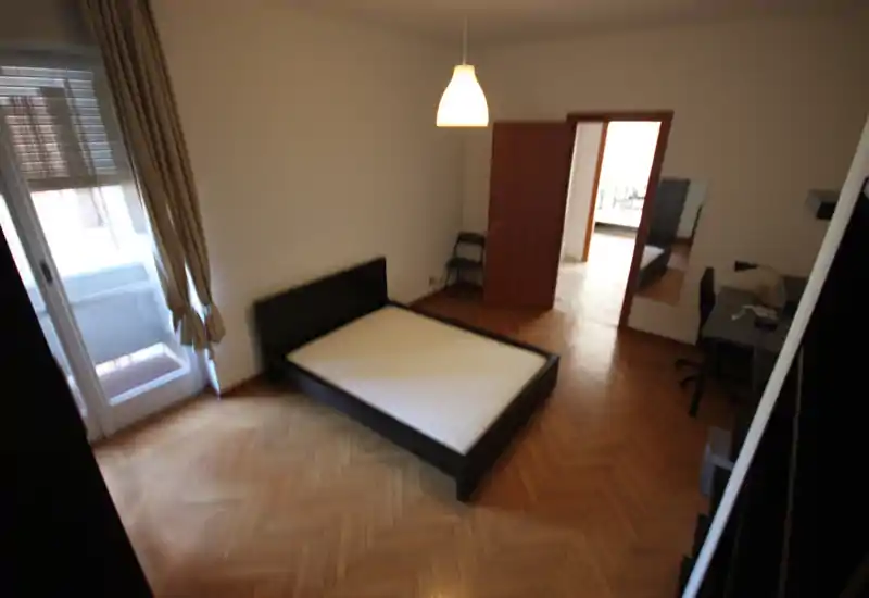 Spacious bedroom with a double bed frame and mattress, parquet floor, desk area and balcony door allowing natural light.