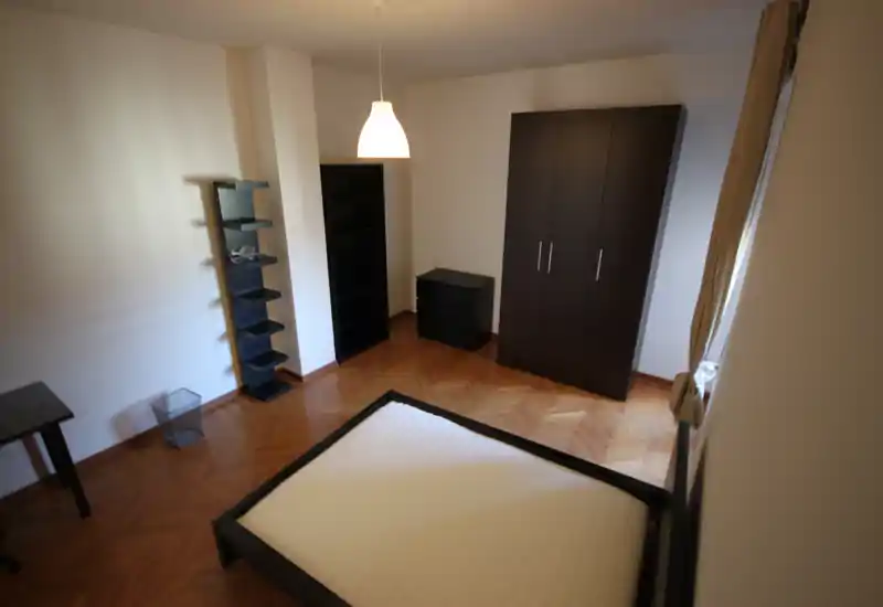 Spacious bedroom with a visible double bed frame, dark wardrobe and matching bedside storage. Natural light from the window highlights the parquet flooring and the room’s clean, minimalist style.