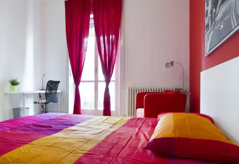Bright, colorful bedroom with a large bed in the foreground, a red armchair, desk and a balcony with sheer curtains letting in natural light.
